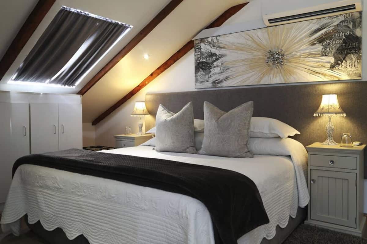 A bedroom with a sloped ceiling, a bed with a white and black bedspread, two bedside tables with lamps, and a large abstract painting on the wall.