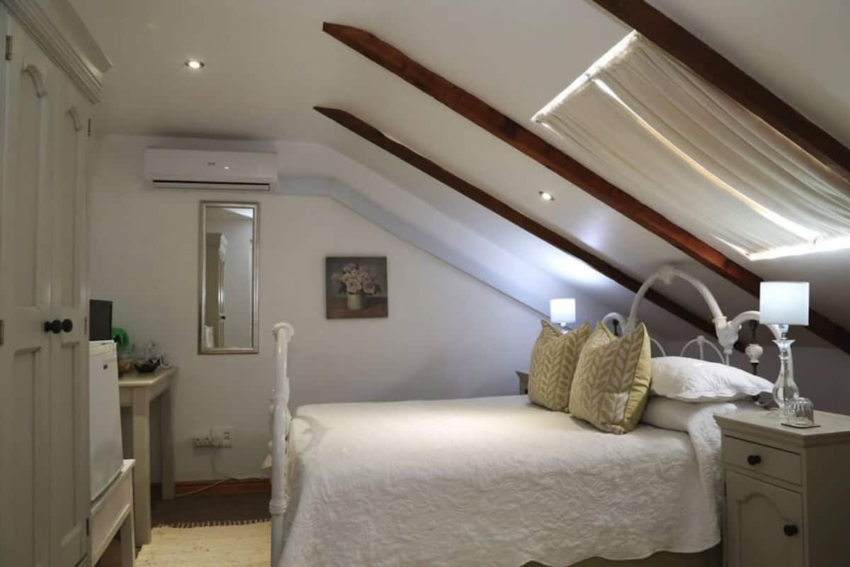 A bedroom with a sloped ceiling, a bed with white bedding, a bedside table with a lamp, and a small bedside table with a glass.
