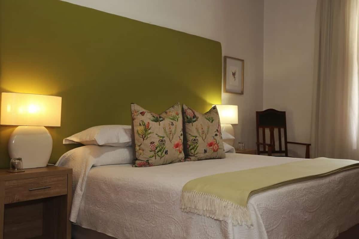 A neatly made bed with floral pillows, a bedside lamp, and a wooden chair in a room with green walls.