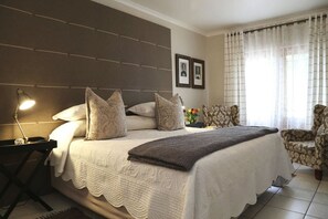 A bedroom with a large bed, a bedside table with a lamp, patterned chairs, and a window with sheer curtains.