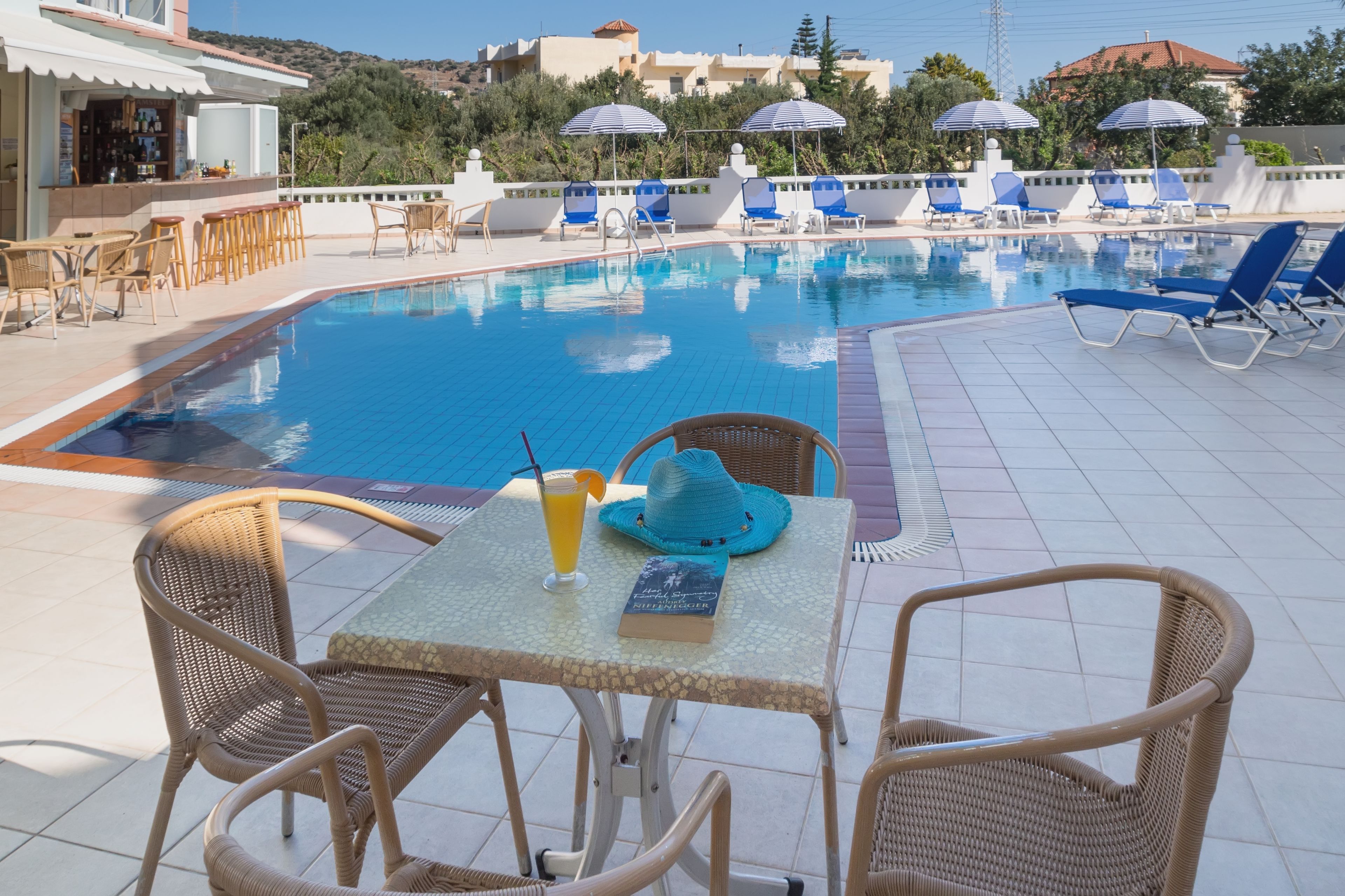Zona de piscina con sillones reclinables, una mesa con una bebida y un libro, y un bar al fondo.