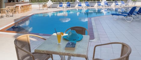 Zona de piscina con sillones reclinables, una mesa con una bebida y un libro, y un bar al fondo.