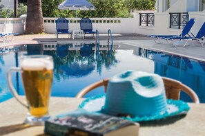 A poolside setting with a clear view of the pool, lounge chairs, and a table with a beverage and a hat. - Athena Apartments (Hersonissos)