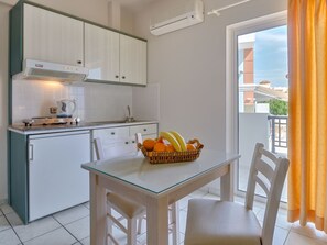 A compact kitchen with white cabinets, a built-in stove, and a dining area with a glass table and chairs. - Athena Apartments (Hersonissos)