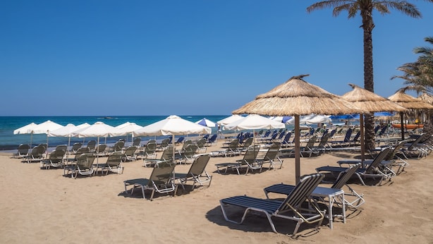 A beach with rows of sun loungers and thatched umbrellas. - Athena Apartments (Hersonissos)
