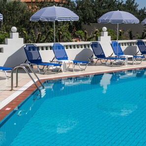A swimming pool with blue and white striped umbrellas, blue lounge chairs, and a tiled deck. - Athena Apartments (Hersonissos)