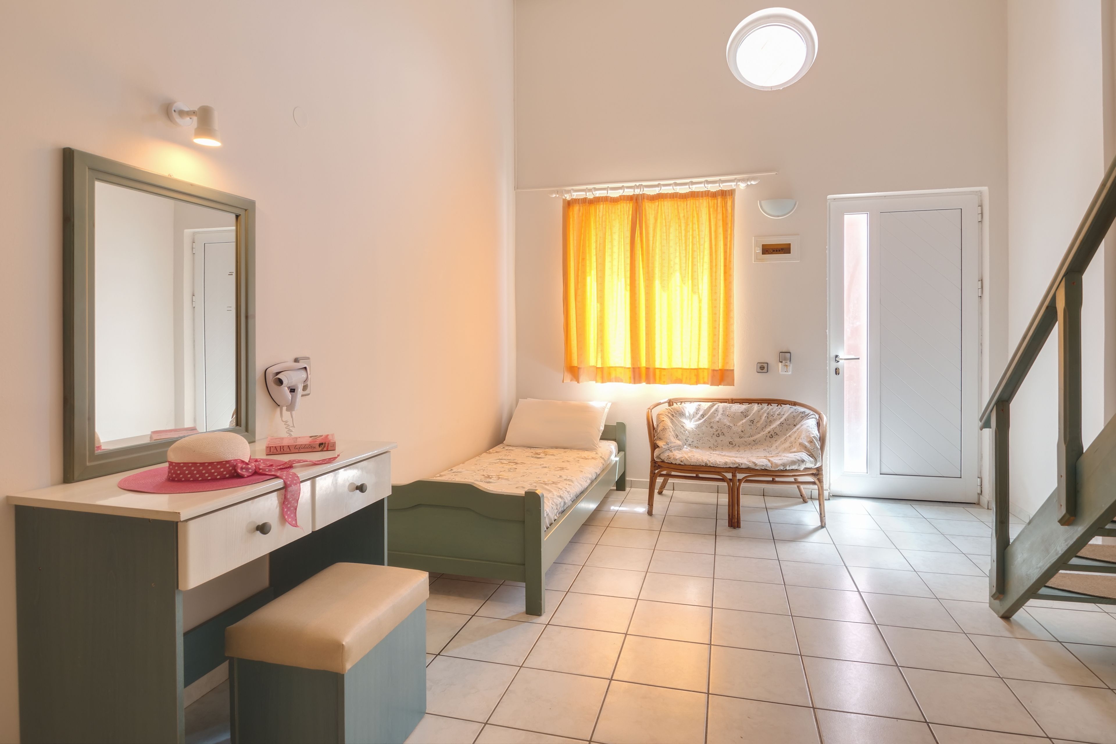 A small, well-lit bedroom with a single bed, a vanity with a mirror, a chair, and a window with curtains.