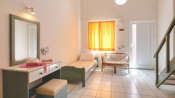 A small, well-lit bedroom with a single bed, a vanity with a mirror, a chair, and a window with curtains.