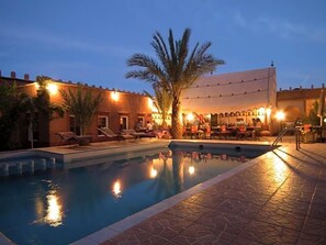 A poolside area with lounge chairs and a bar, surrounded by buildings with warm lighting. - Tomboctou (Tinghir)
