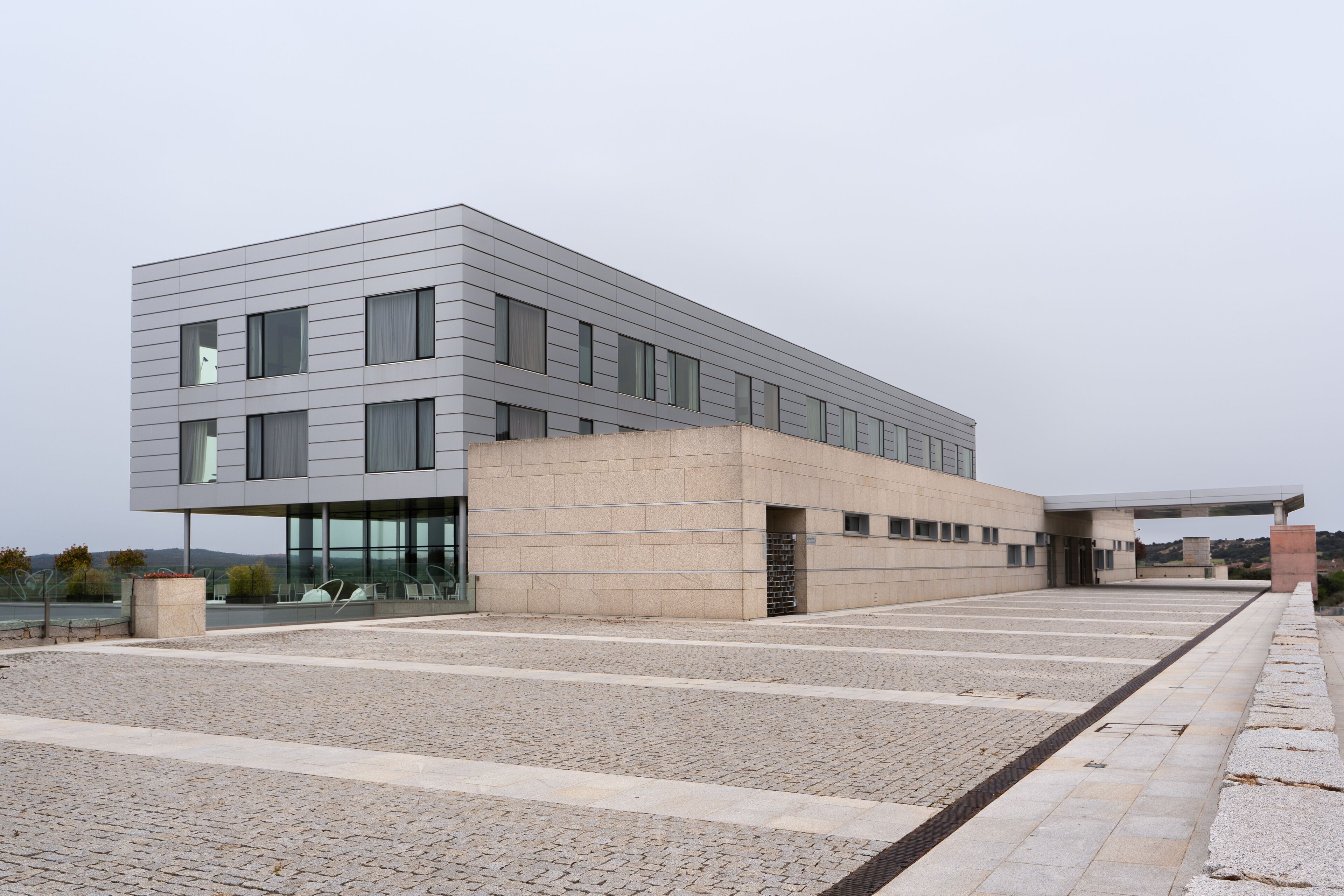 Un edifici modern amb teulada plana, grans finestres i un pati empedrat.