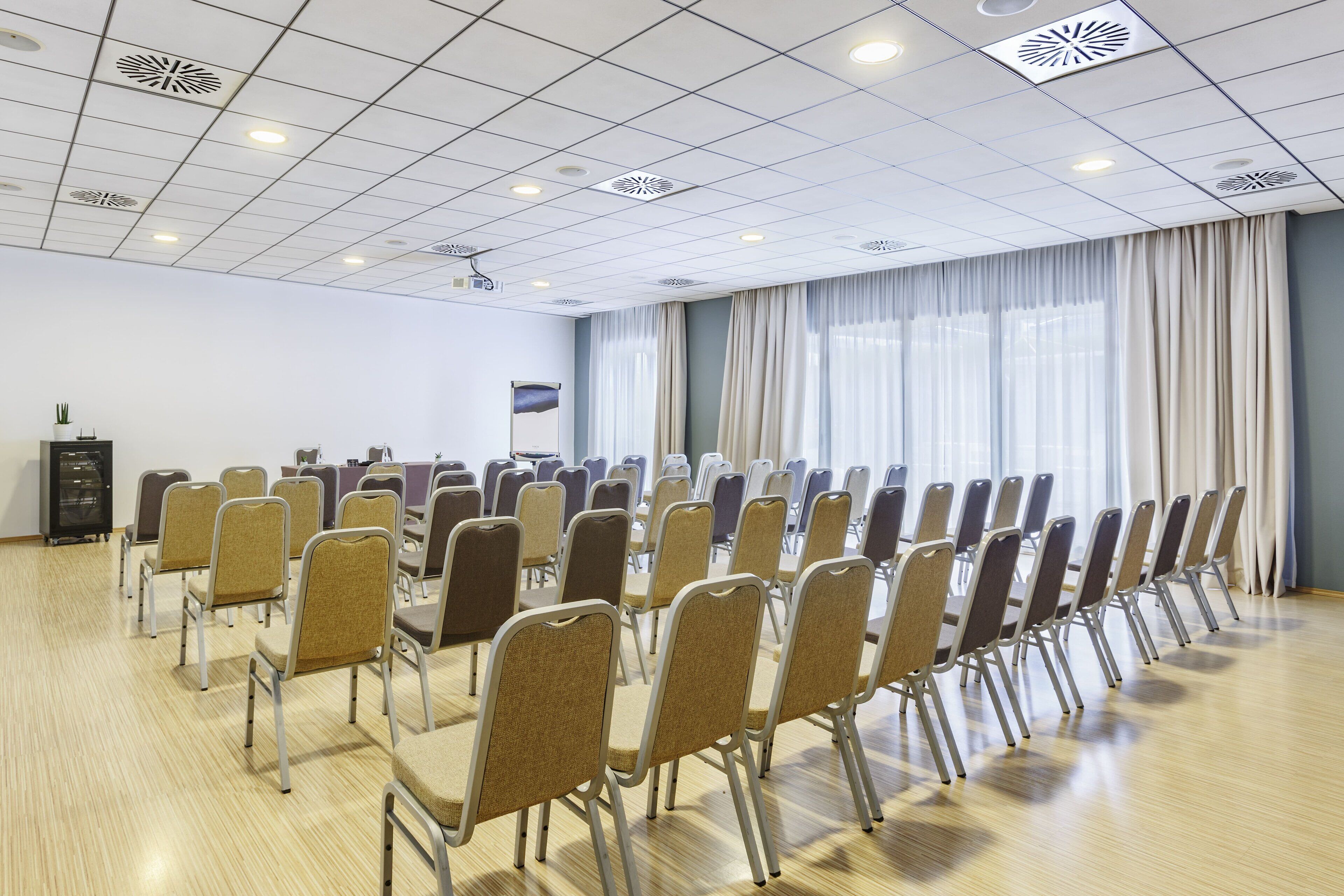 Uma sala de conferências com fileiras de cadeiras, uma tela de projetor e uma grande janela com cortinas.