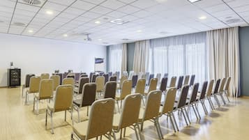 Uma sala de conferências com fileiras de cadeiras, uma tela de projetor e uma grande janela com cortinas.