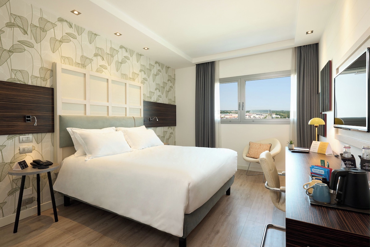 Une chambre d’hôtel avec un grand lit, un bureau avec une machine à café, une chaise et une vue sur la ville.