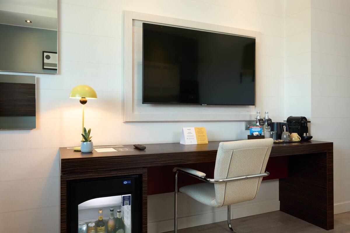 Een moderne hotelkamer met een houten bureau, een comfortabele stoel, een flatscreen televisie, een koffiezetapparaat en een minibar.