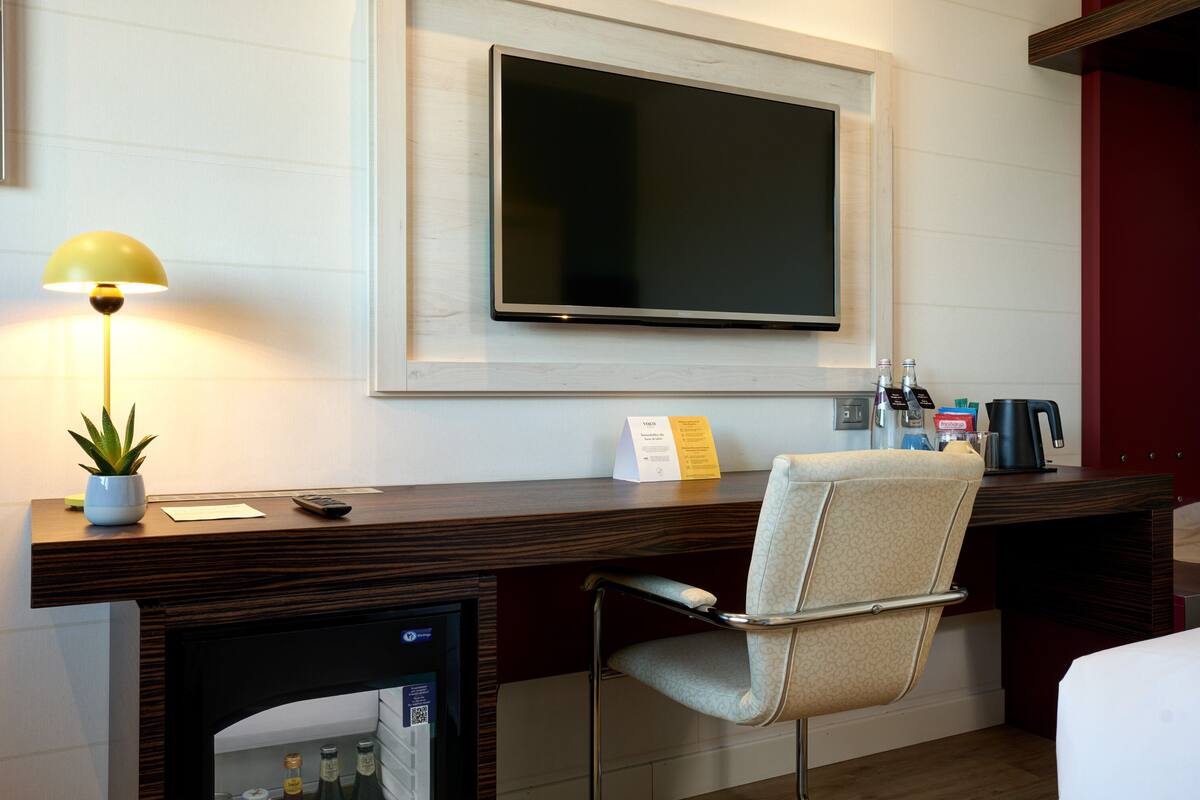 Een hotelkamer met een bureau, een stoel, een aan de muur bevestigde televisie, een lamp, een kleine potplant en een minibar.