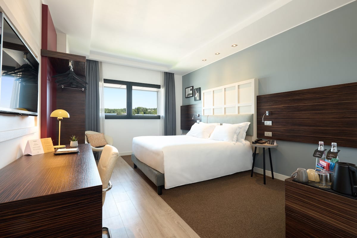Une chambre d’hôtel avec un lit, un bureau, une chaise et une petite table sur laquelle se trouvent une lampe et une plante.