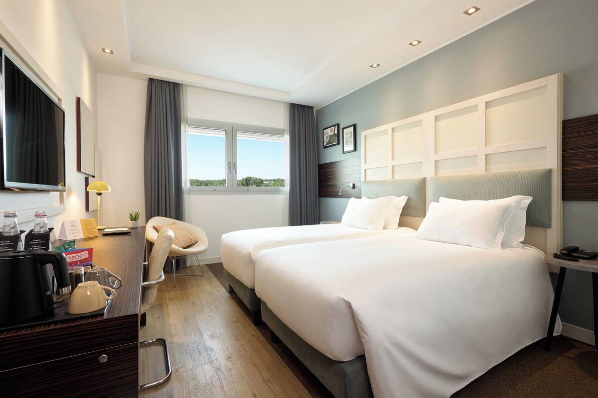 Een moderne hotelkamer met twee bedden, een bureau met koffiezetapparaat, een televisie en een raam met uitzicht.