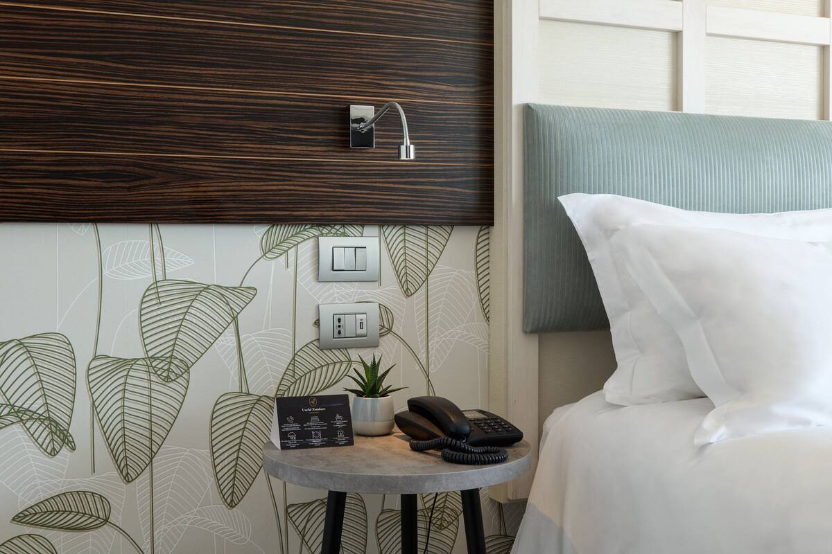 Hotelkamer met een bed, nachtkastje met telefoon, kleine plant en een lichtschakelaar aan de muur.