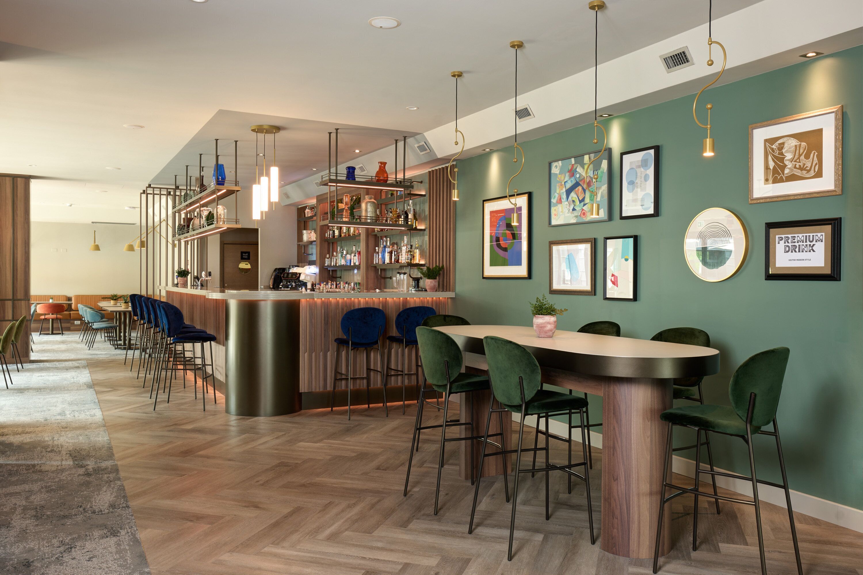 A bar area with a wooden bar counter, green bar stools, and a variety of bottles and glasses. There are framed artworks on the walls and a potted plant on the counter.