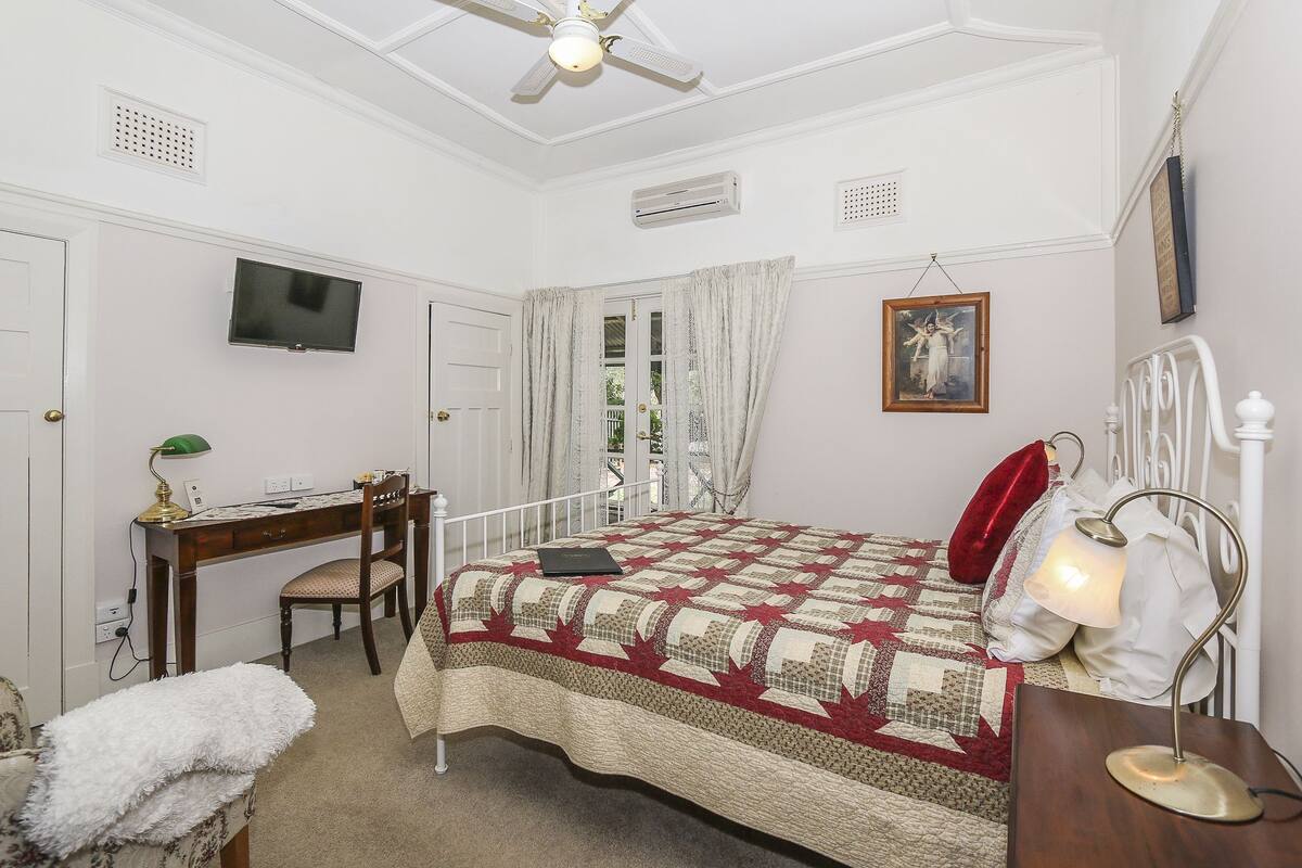 Um quarto com cama, escrivaninha, cadeira, luminária e televisão.