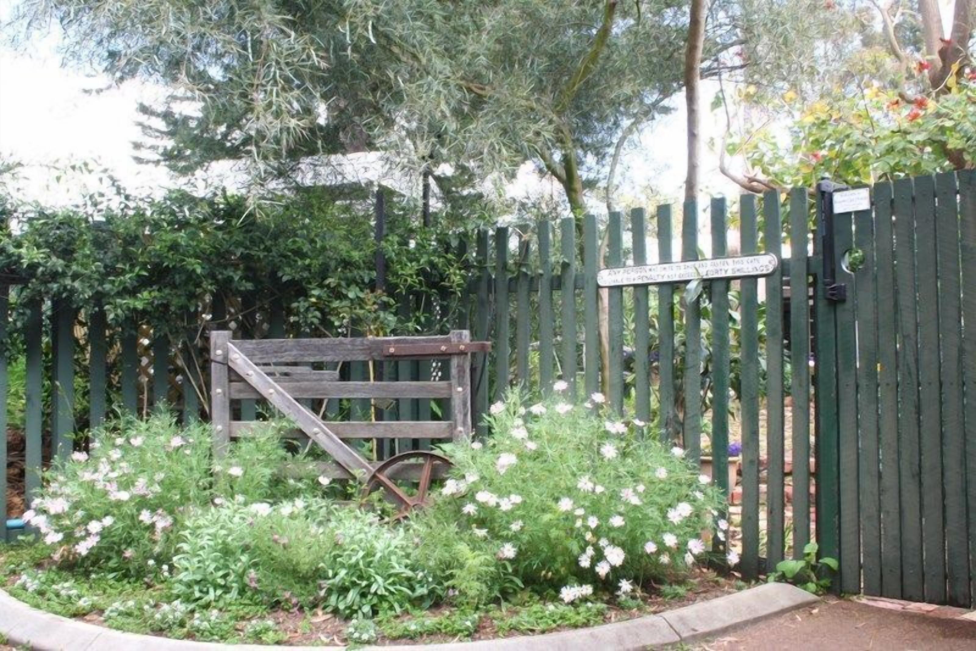 Une porte en bois, agrémentée d’une pancarte, est entourée d’un jardin où poussent différentes plantes et fleurs.