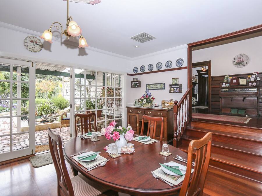 Comedor con mesa de madera dispuesta para seis personas, rodeada de sillas. Hay un ventanal con vistas a un jardín. Una escalera conduce a un segundo piso donde se ve una cocina.