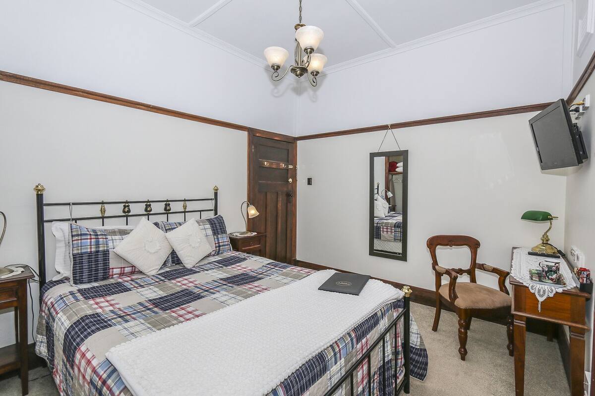 Quarto com cama, escrivaninha, cadeira, televisão e espelho.