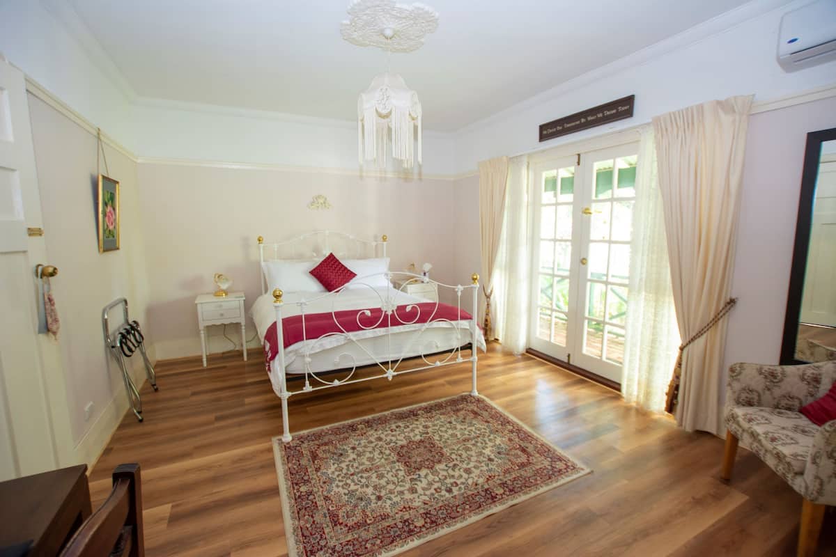 Quarto com piso de madeira, cama branca com travesseiro vermelho, mesa de cabeceira, cadeira e uma janela grande com cortinas.