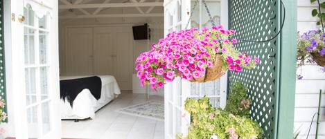 Uma porta aberta dando acesso a um quarto com uma cama, um cesto de flores pendurado e um vaso com planta.