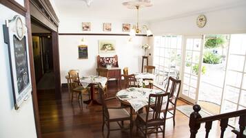 A dining area with wooden tables and chairs, a chandelier, and a view of the garden through large windows.