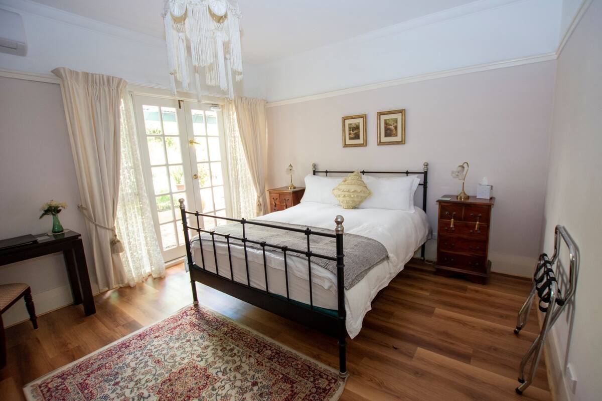 Quarto com cama, mesas de cabeceira, poltrona, escrivaninha, janela com cortinas e lustre.