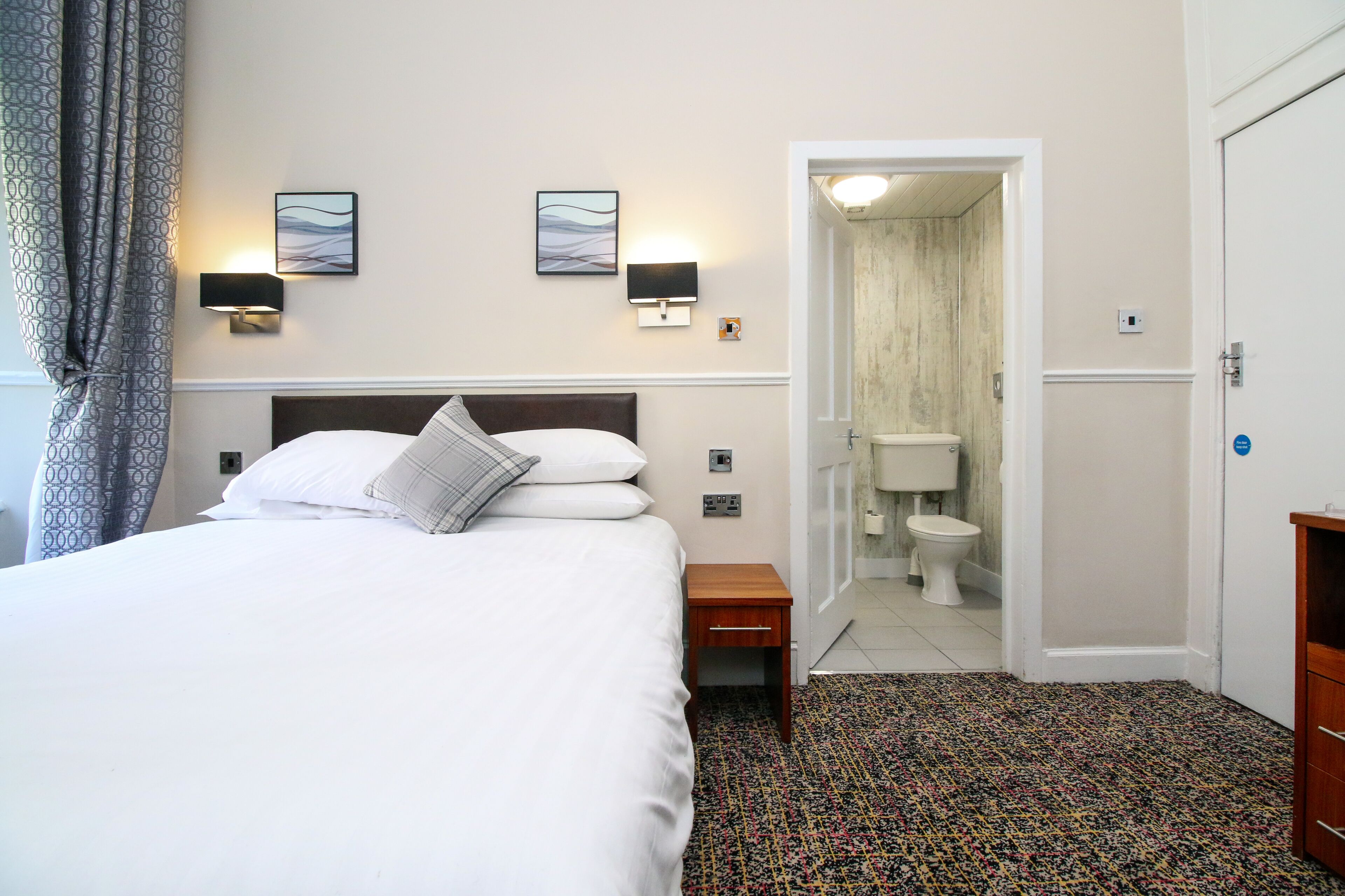 A hotel room with a bed, two wall-mounted lamps, a small wooden nightstand, a patterned carpet, and a bathroom visible through an open door.