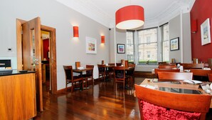 Een restaurantruimte in een hotel met houten meubels, een rode fauteuil en een groot raam met jaloezieën.