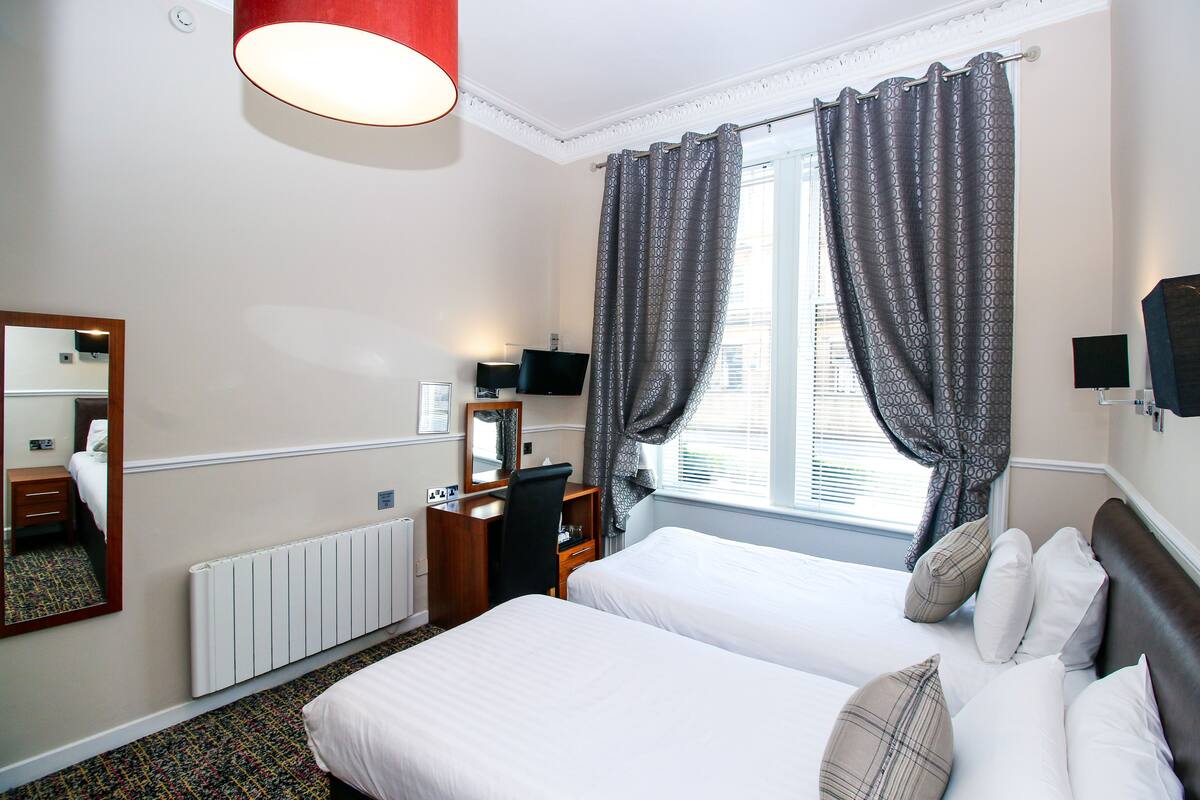 A hotel room with two beds, a desk, a chair, a mirror, a TV, and a window with curtains.