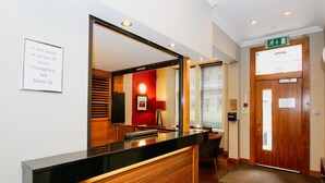 Een hotel receptie met een houten balie, een bordje met Kamer 15 voor noodgevallen, en een deur met een nooduitgangsbord.