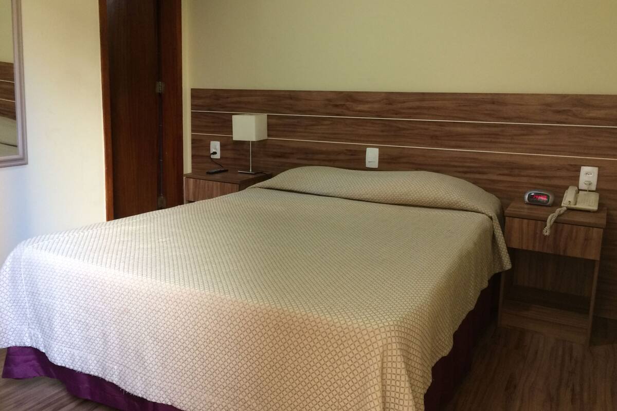 A hotel room with a bed, a nightstand with a phone and a lamp, and a wooden headboard.