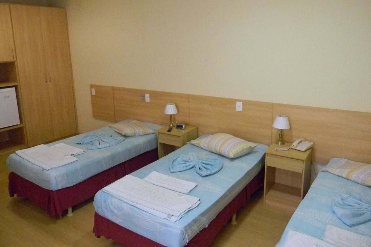 A hotel room with three single beds, each with a pillow and a bedside lamp. There is a small table with a telephone and a lamp. The room has wooden cabinets and a white ceiling light.