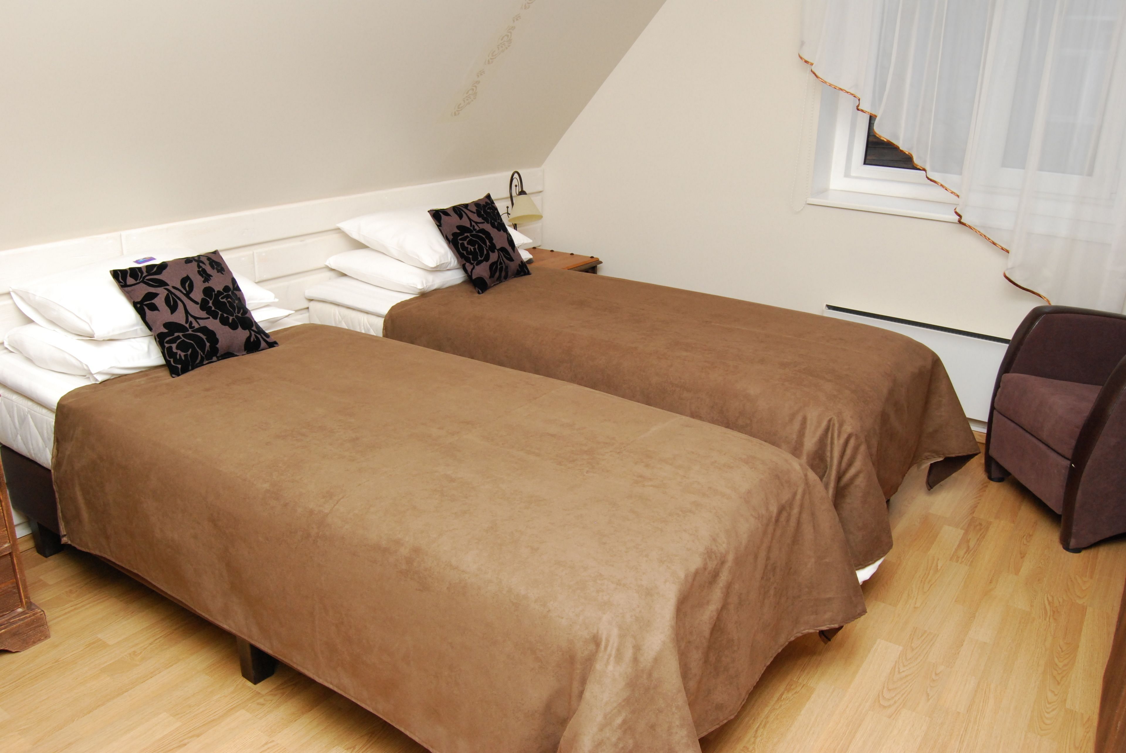 A room with two single beds, each with a brown bedspread and a black floral pillow. There is a wooden nightstand with a lamp between the beds.