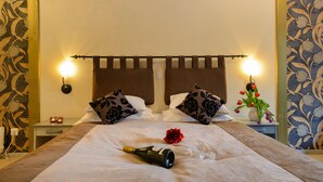 A neatly made bed with a bottle, glass, and rose on the bedspread, two pillows with a floral pattern, a bedside table with a candle, and a wall-mounted light fixture. - Hotel Hansalinn (Parnu)