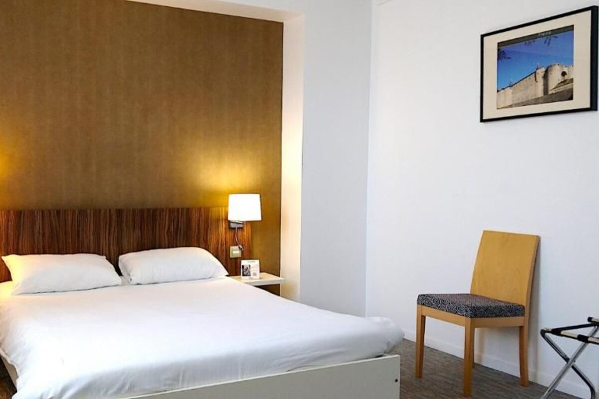 A hotel room with a bed, two bedside lamps, a chair, and a framed picture on the wall.