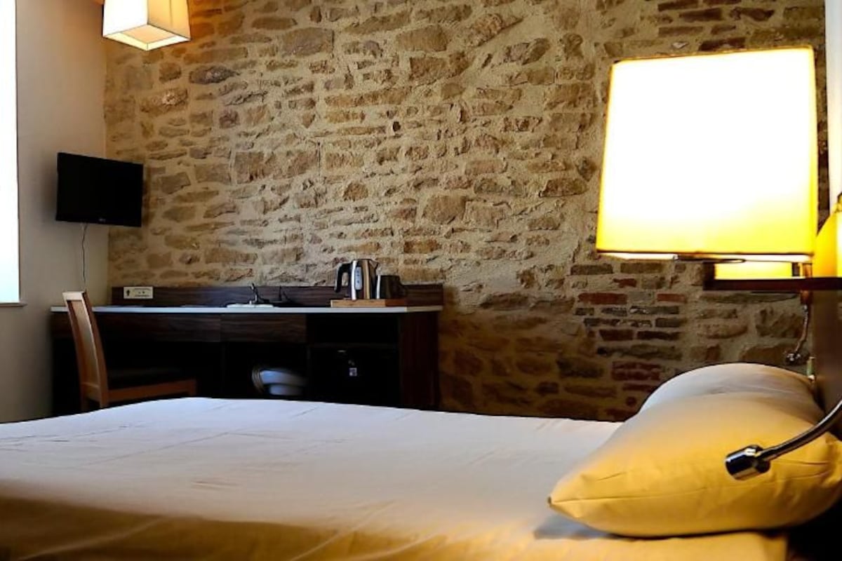 Habitación de hotel con pared de piedra, una cama con almohada, un escritorio con silla, un televisor y una lámpara.