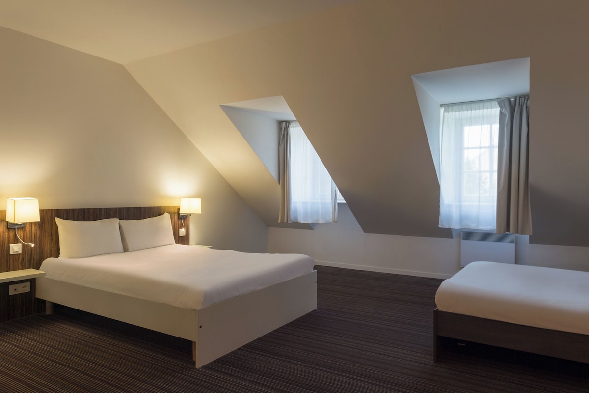 A hotel room with a bed, a bedside table with a lamp, a window with curtains, and a radiator.