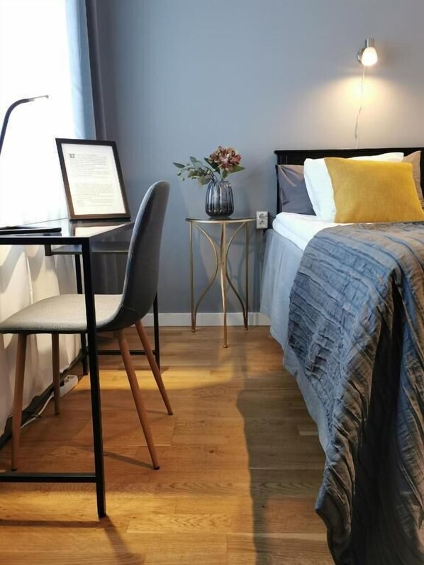 Una habitación de hotel con cama, escritorio con silla, una mesita con un florero y una lámpara de pared.
