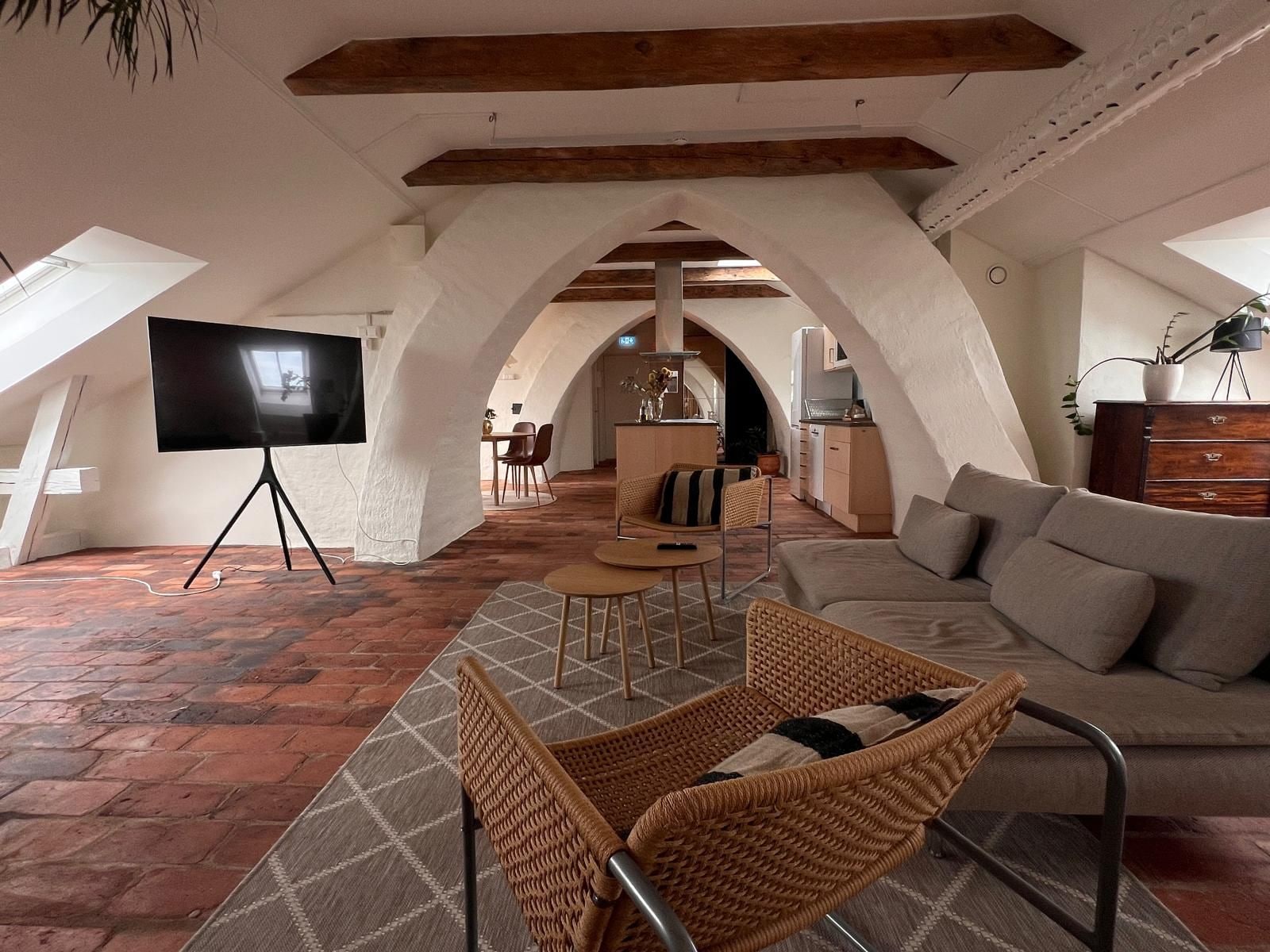 An interior with a TV on a tripod, a sofa, a wicker chair, and a kitchen in the background.
