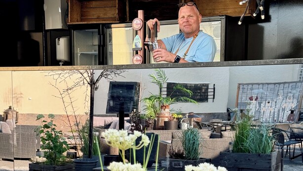 Una persona de pie en un área de estar exterior moderna con mesas y sillas de madera, rodeada de plantas en macetas y una terraza de madera.