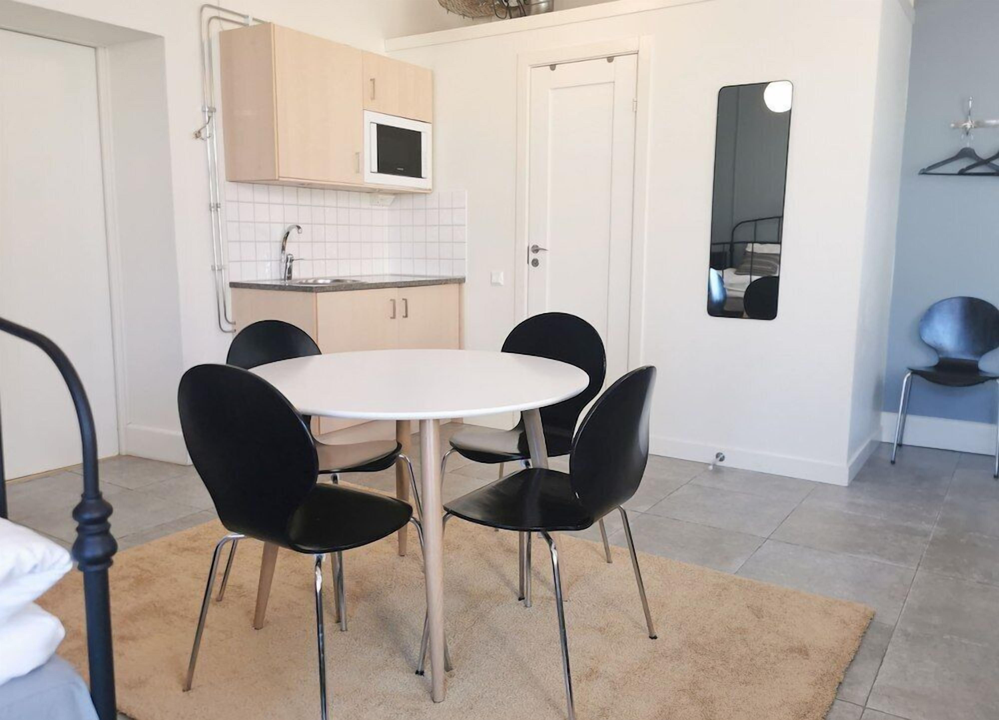 Une cuisine compacte avec un plan de travail blanc, un four à micro-ondes et un évier. On y trouve une table ronde avec quatre chaises et un tapis beige sur le sol.