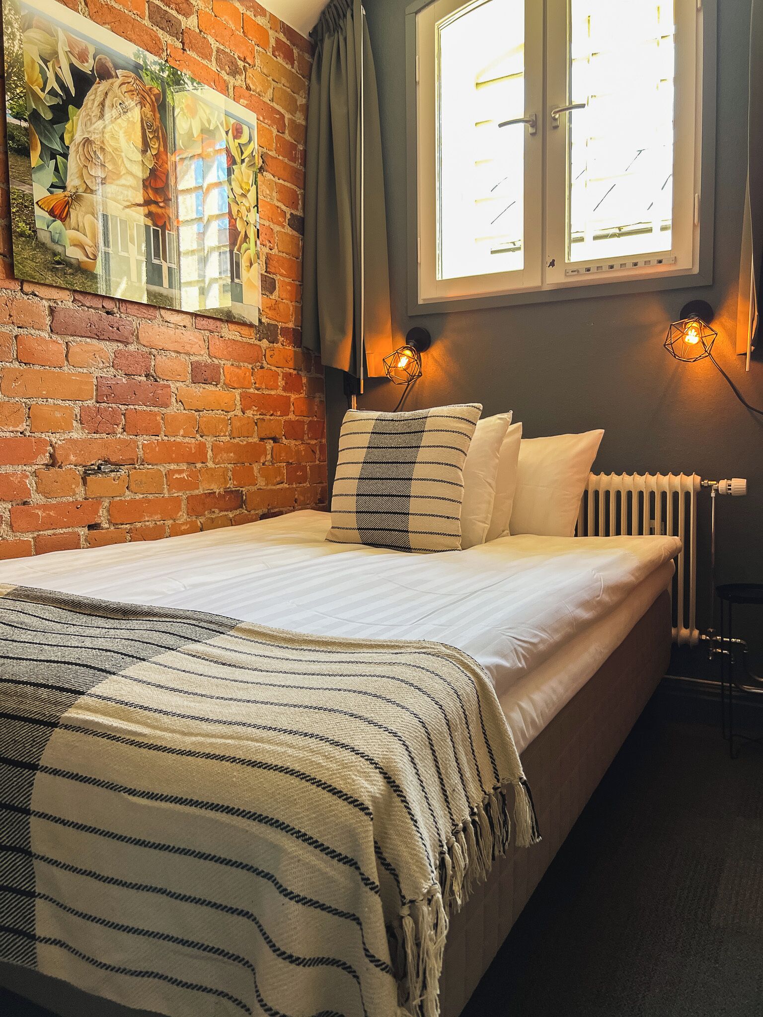 A bedroom with a brick wall, a large window, a bed with white bedding, striped pillows, and a throw blanket.