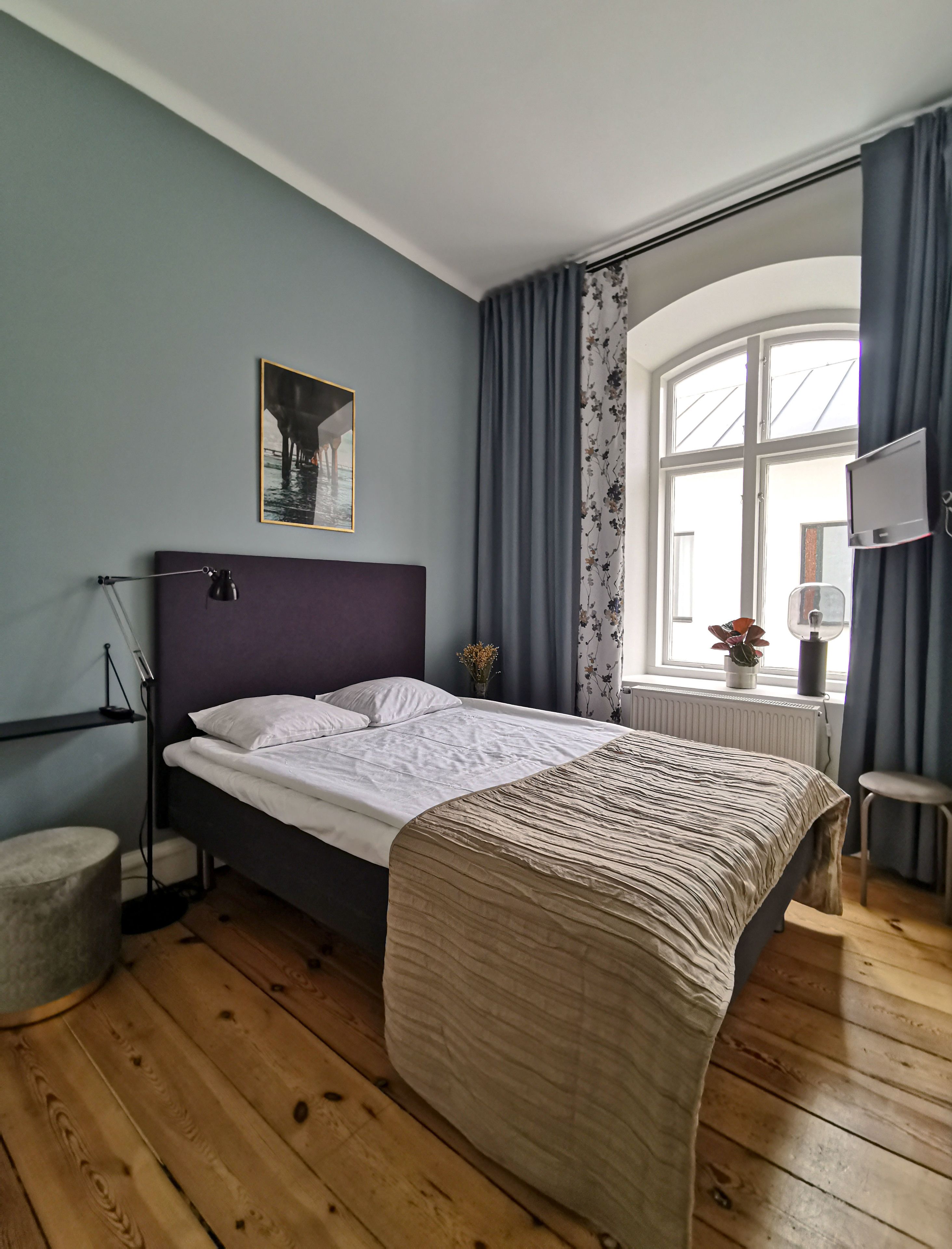 A bedroom with a bed, a desk, a chair, a window with curtains, a radiator, and a wall-mounted TV.