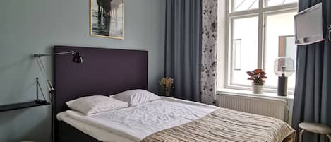 A bedroom with a bed, a desk, a chair, a window with curtains, a radiator, and a wall-mounted TV.
