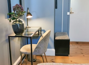 Una habitación con una mesa de vidrio, una silla, un pequeño banco, un florero con flores y una lámpara.